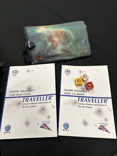 A photograph with a dice bag at the top (which has a galaxy image) and then two copies of BITS Double Adventure 1 'Delta 3 is Down' / 'Cold Dark Grave', for Traveller. The cover is white with an inverted image of stars. The following logos are shown: Top left - the TAS logo, bottom left - the BITS logo and bottom right the TravCon Scoutship with the year beside it. The Traveller logo and line across the cover are dark blue. There are three Traveller six-sided dice lying on it.