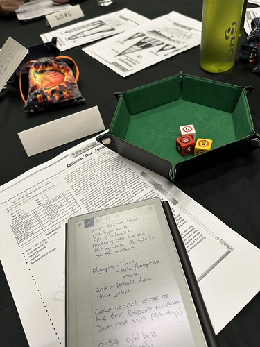 TravCon 2025 - midsession photo showing notes on a reMarkable Move, the character sheet and a dice tray lined with green felt with dice in it. In the centre of the table is the GM's BITS dice bag and some details about the ship we were hunting.