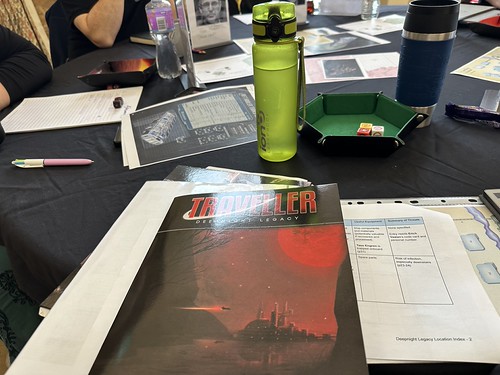 Table photo at TravCon 2025. Deepnight Legacy lies in front of me, with a table summarising the plot part hidden underneath the book (with columns for Useful Equipment and Summary of Threats peeking out). The centre of the table has drinks, dice, maps and starship diagrams.